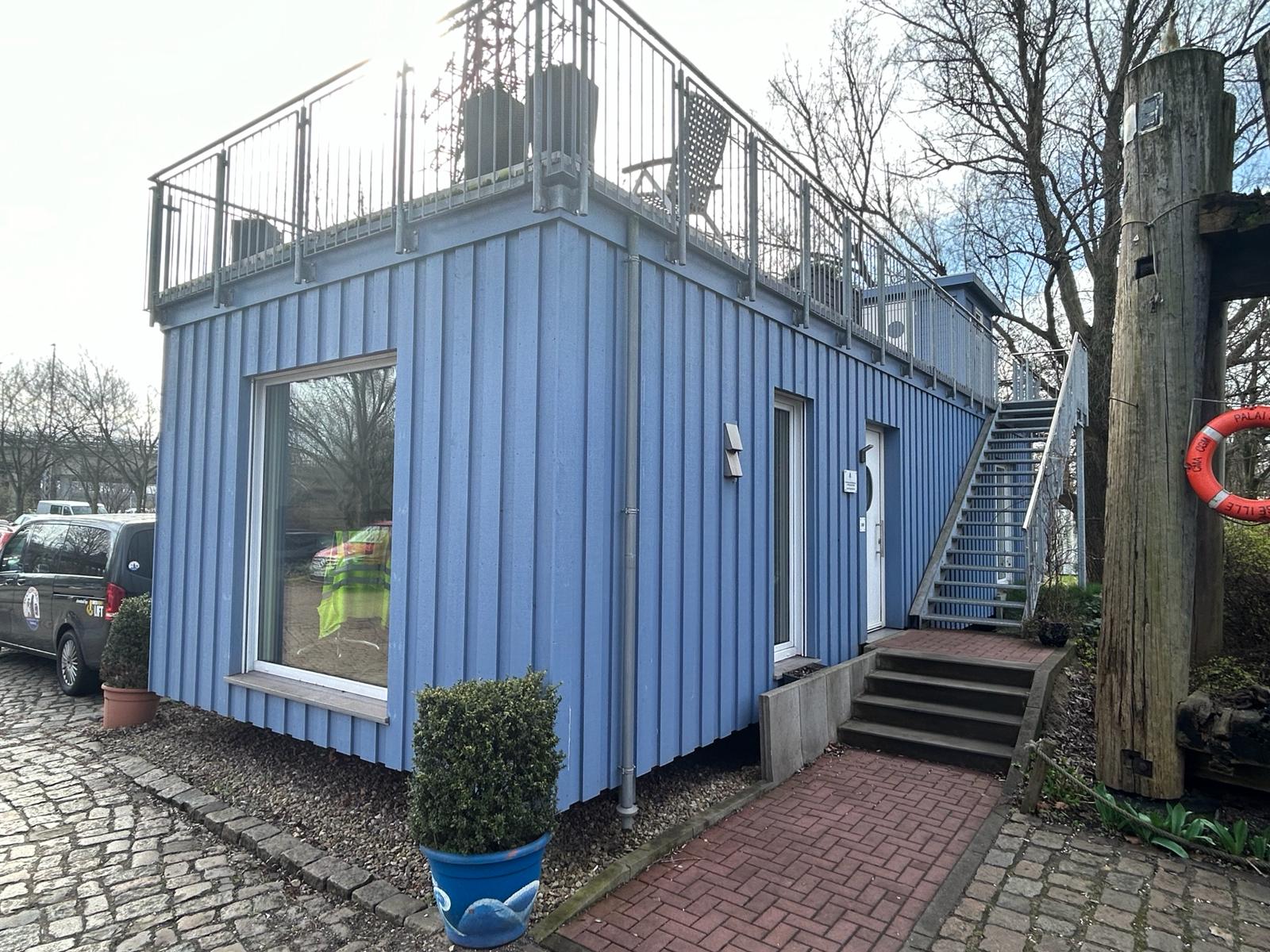 A smallish blue prefabricated office building. The building is single storey with steps up to a balcony garden on the flat roof with plants and garden furniture. 